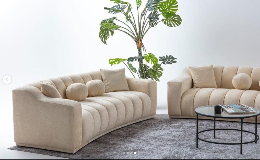 Beżowa, tapicerowana sofa o nowoczesnym, falistym kształcie z pionowymi przeszyciami.